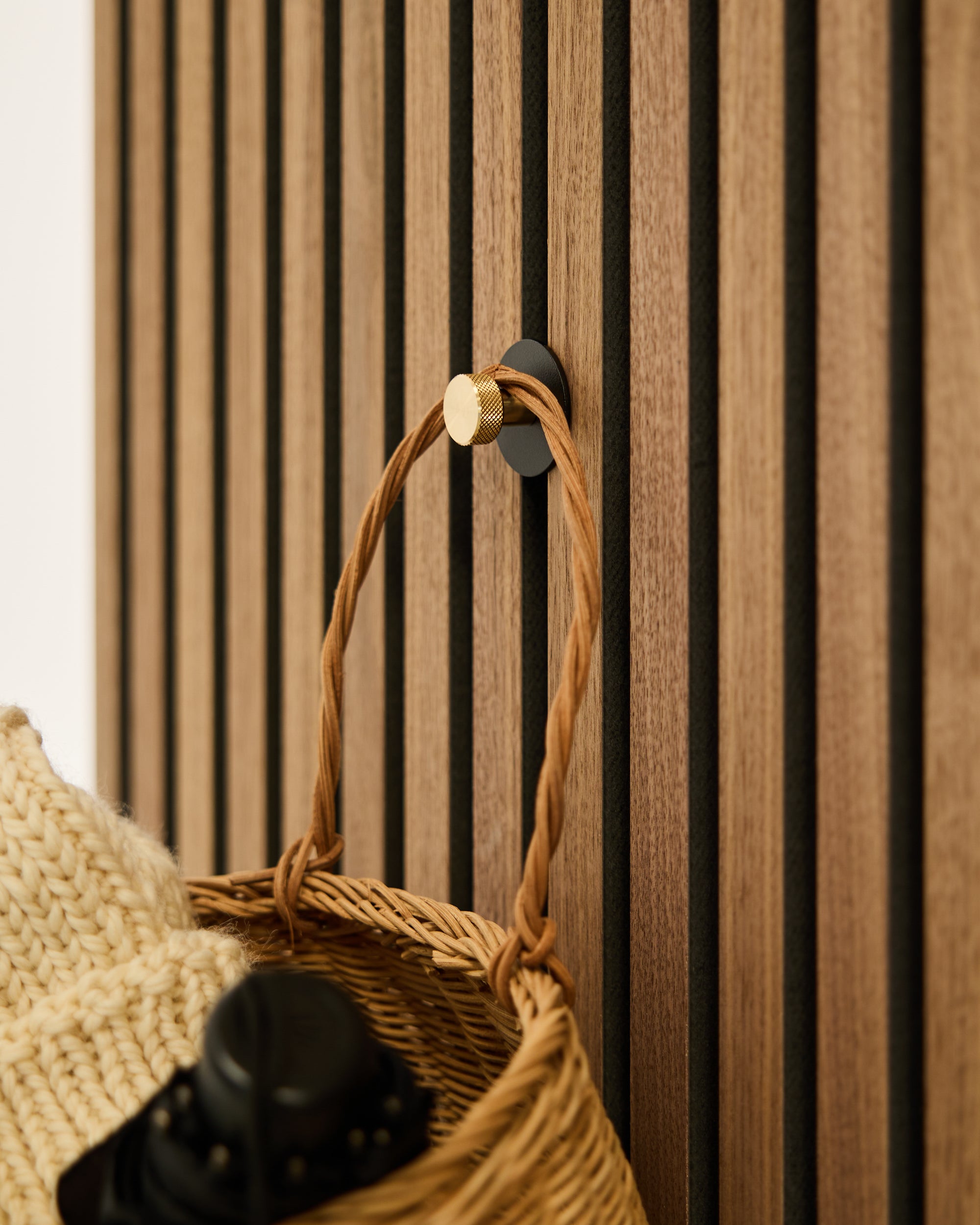 A woven basket with a braided handle hangs on a round gold wall hook, adding texture to a vertical Nordic acoustic slatwall panel.
